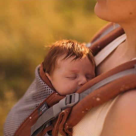 Mãe sentada num campo verdejante ao pôr do sol, usando a Mochila Love and Carry ONE com o bebé.