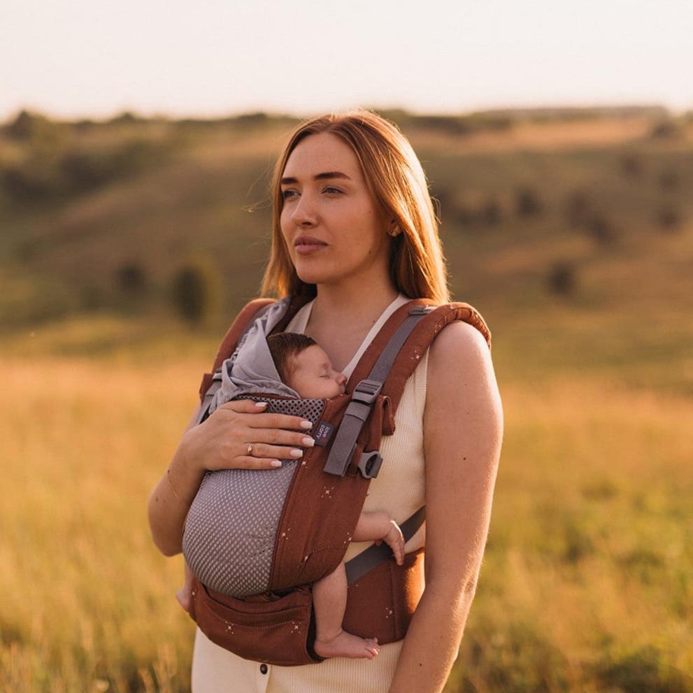 Mãe de pé num campo, com a Mochila Love and Carry ONE, segurando o bebé em posição de transporte frontal.