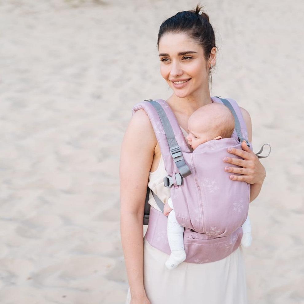 Mulher sorridente usando a mochila Love & Carry ONE - Bloom, segurando um bebé no peito, num ambiente de praia.