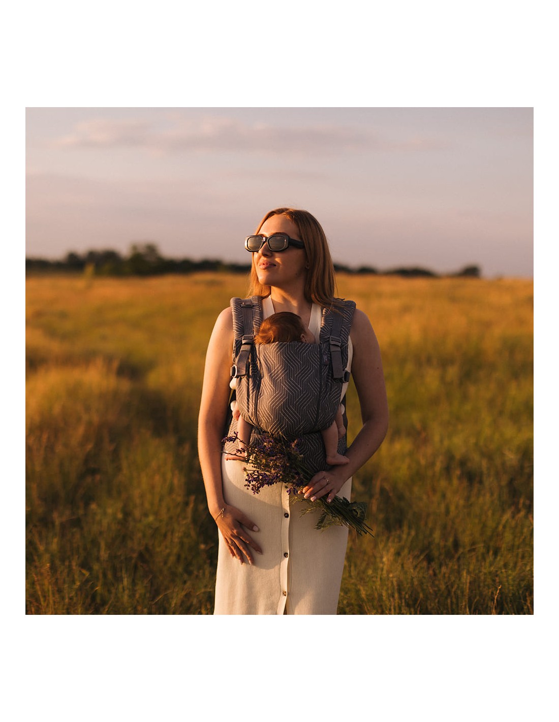 Mãe a caminhar num campo com o bebé na mochila, ao pôr do sol.