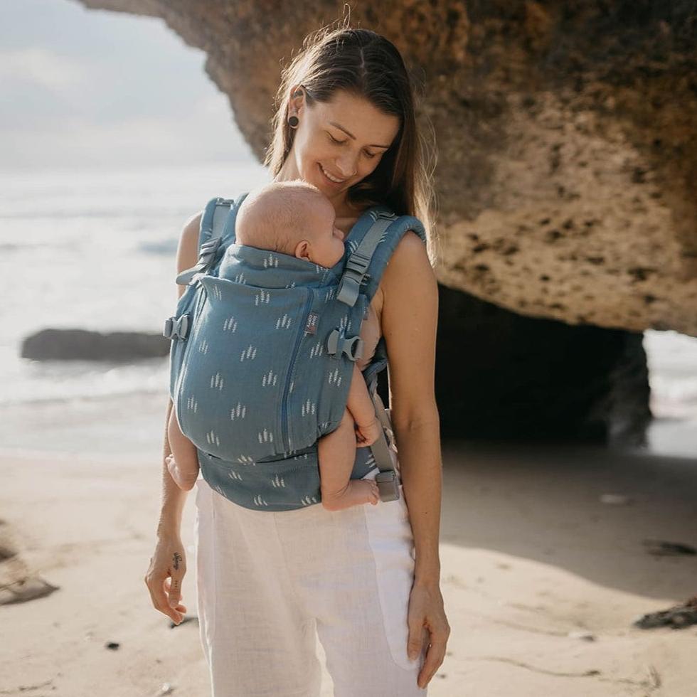 Mulher a caminhar na praia usando a Mochila Love and Carry ONE, com o bebé a dormir dentro da mochila.