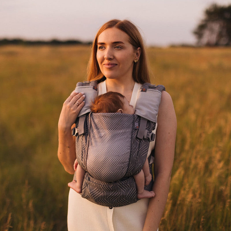 Mãe sorridente a usar porta-bebés com protetores de alças cinzentos, num cenário exterior ao pôr do sol.