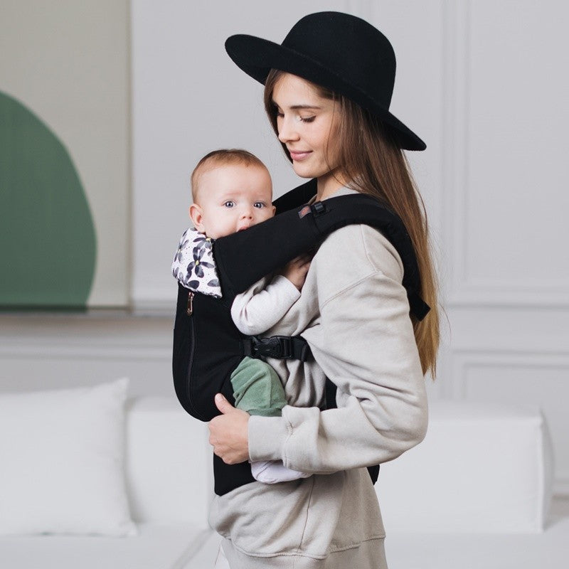 Mãe com chapéu preto a usar um porta-bebés ergonómico preto, focando-se nos protetores de alças pretos Love and Carry no ombro.