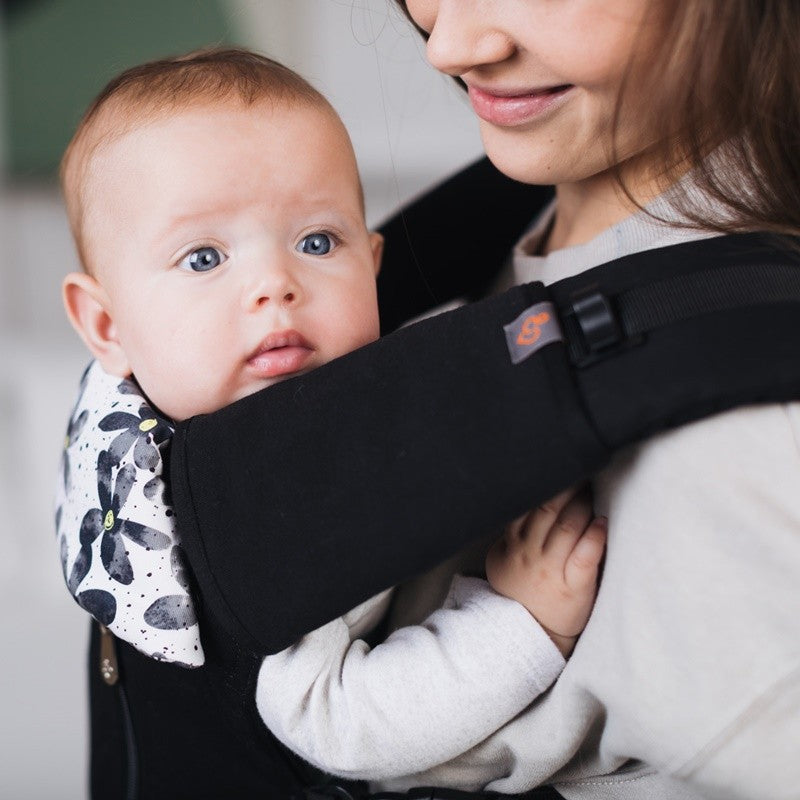 Bebé de olhos azuis no porta-bebés preto, com protetores de alças pretos e um forro estampado floral, em contacto visual com a mãe.