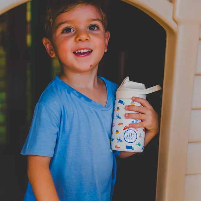 Criança sorridente, de cabelo castanho e t-shirt azul, a segurar a garrafa térmica Cool Bottles Little Wheels de 350ml com padrão de veículos.