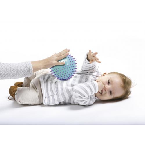 Bebé deitado a brincar com uma bola sensorial azul-claro, enquanto uma mão adulta a guia, estimulando o desenvolvimento.