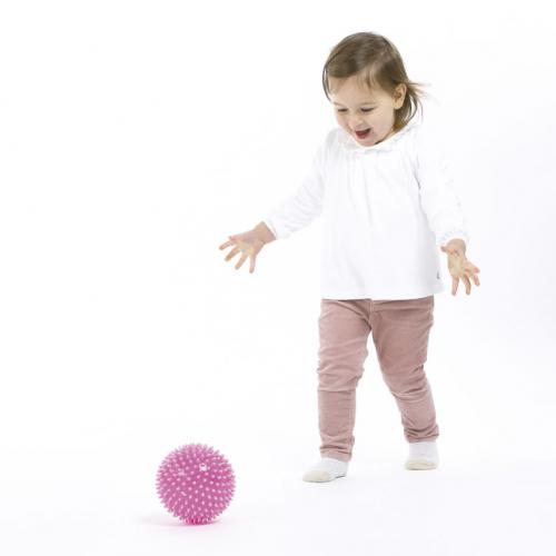 Criança pequena a sorrir e a brincar com uma bola sensorial rosa com relevos no chão, promovendo a coordenação.