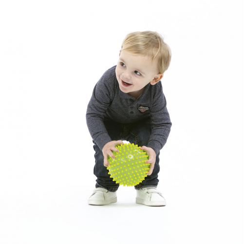 Criança pequena sorridente a segurar uma bola sensorial verde-lima com relevos, estimulando o tato.