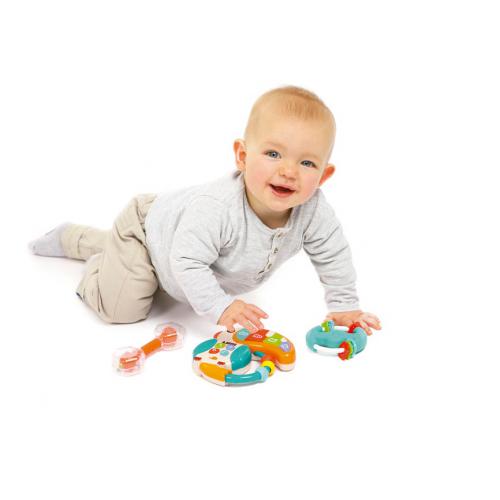 Bebé sorridente a gatinhar, rodeado pelos três brinquedos musicais Ludi: tucano, maraca e anel pandeireta, em fundo branco.