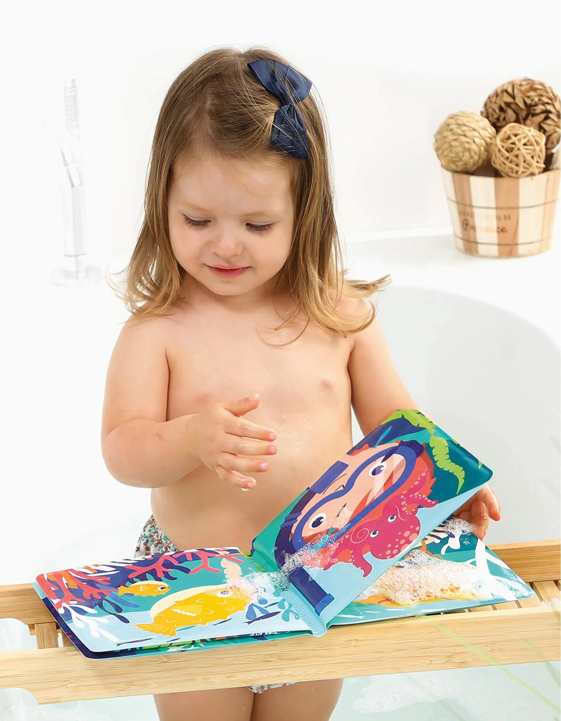 Menina pequena sorridente a segurar o Livro de Banho Mágico Ludi aberto, com ilustrações coloridas de animais marinhos e um polvo.
