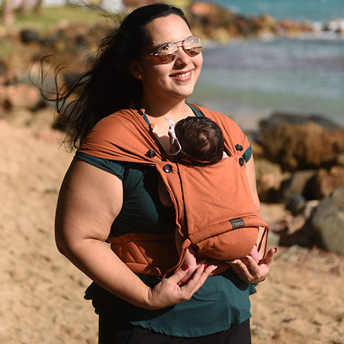 Mãe sorridente a carregar bebé recém-nascido numa mochila de babywearing Neko Tiny cor terracota na praia, mostrando ajuste ergonómico.