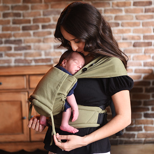 Mãe a beijar a cabeça do bebé recém-nascido que dorme na mochila de babywearing Neko Tiny cor verde azeitona, em ambiente interior.