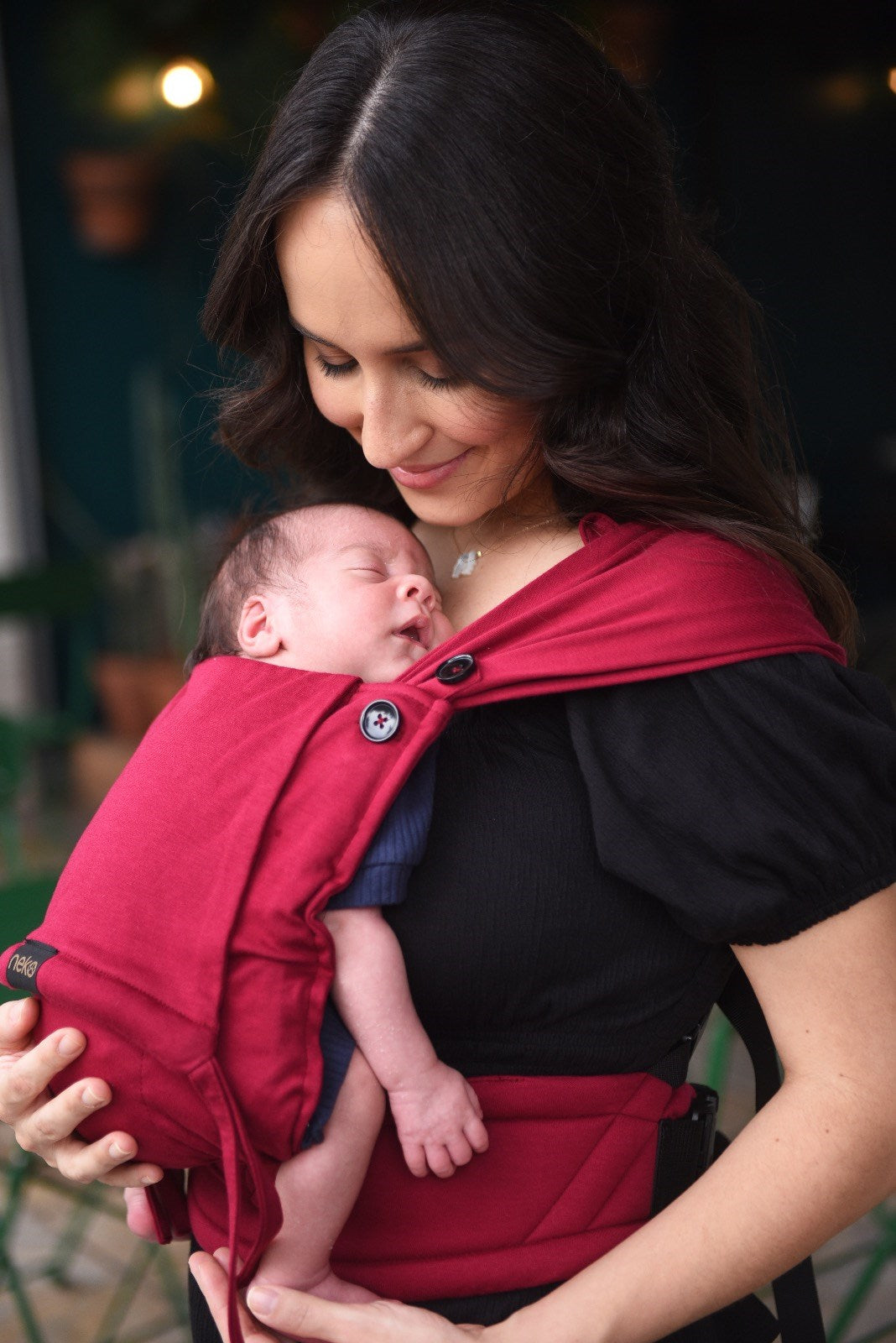 Mãe a olhar para o bebé recém-nascido na mochila de babywearing Neko Tiny cor vermelha, mostrando o ajuste e conforto.