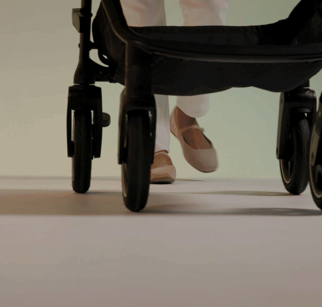 Mãe sorridente a passear com o seu bebé no carrinho Joie versadrift preto num passeio urbano com vegetação.