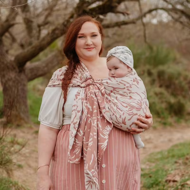 Mãe com cabelo ruivo a usar o Sling Neobulle SEA CELL beije rose para transportar o bebé, em ambiente exterior com árvores.