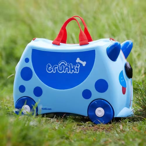 Mala de viagem Trunki azul com design de dinossauro pousada sobre relva verde, mostrando o seu aspeto exterior.
