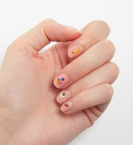 Mão com unhas curtas decoradas com autocolantes OMY de comida, incluindo um abacate, um foguetão e uma banana.