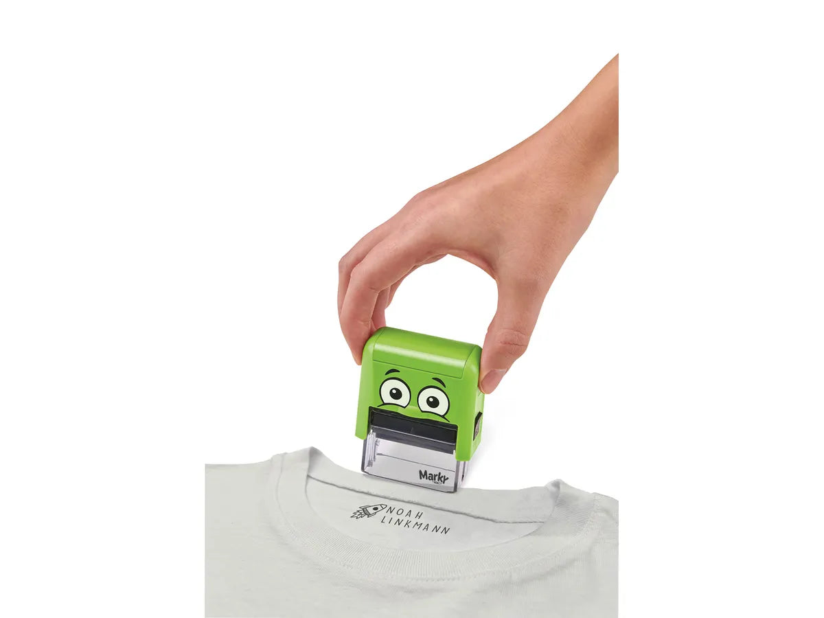 Demonstração do uso do carimbo MARKY Mine DIY a aplicar um nome ("NOAH LINKMANN") na etiqueta de uma t-shirt branca.