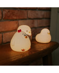 Luz noturna em forma de galinha de silicone branca, com detalhes em laranja nas patas e crista vermelha, iluminada com luz branca quente, ao lado de outras luzes noturnas em forma de animal.