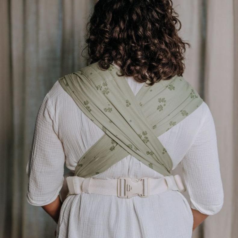 Vista traseira de uma pessoa com cabelo encaracolado a usar o Mei Tai Fidella FlyClick Toddler na cor verde claro com padrão de trevos.