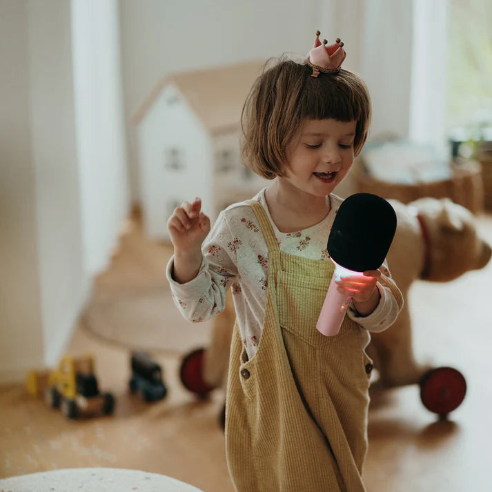 Menina pequena com coroa a cantar alegremente num microfone de karaoke KidyWolf rosa com luzes LED.