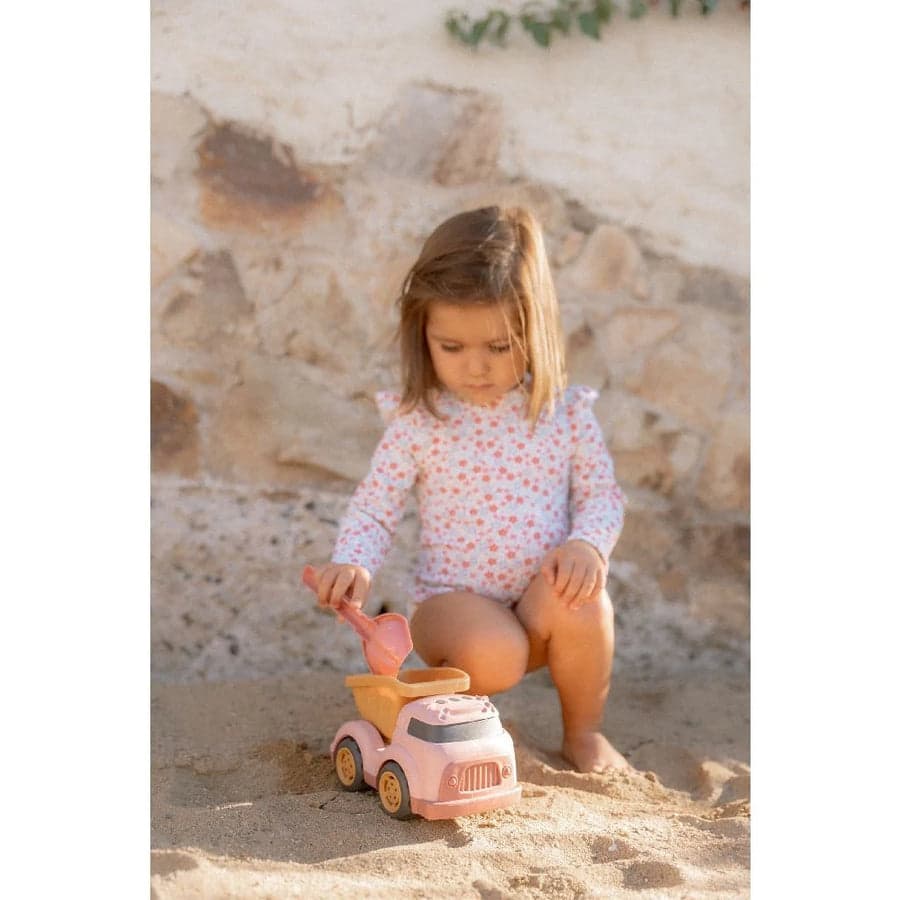 Menina pequena, de fato de banho estampado, a brincar com o camião de reboque Little Dutch na areia junto a uma parede de pedra.
