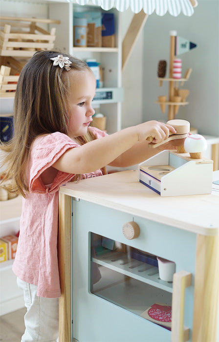 Uma menina brinca com a gelataria compacta de madeira small foot, servindo bolas de gelado com uma colher de madeira.