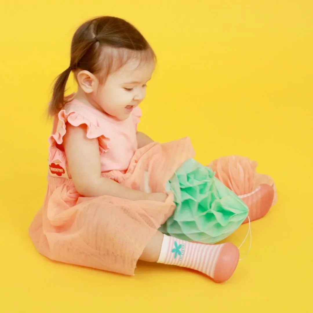 Menina a sorrir usando Attipas Peekaboo Peach, calçado barefoot rosa e branco com flor turquesa, destacando-se em fundo amarelo.