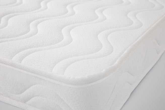 Detalhe do canto de um colchão viscoelástico Micuna branco para berço, mostrando a costura do ribete e o padrão ondulado do tecido acolchoado.
