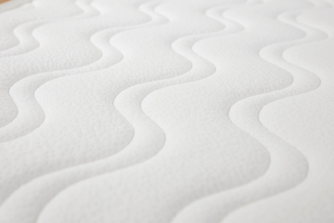 Detalhe do canto de um colchão viscoelástico Micuna branco para berço, mostrando a costura do ribete e o padrão ondulado do tecido acolchoado.