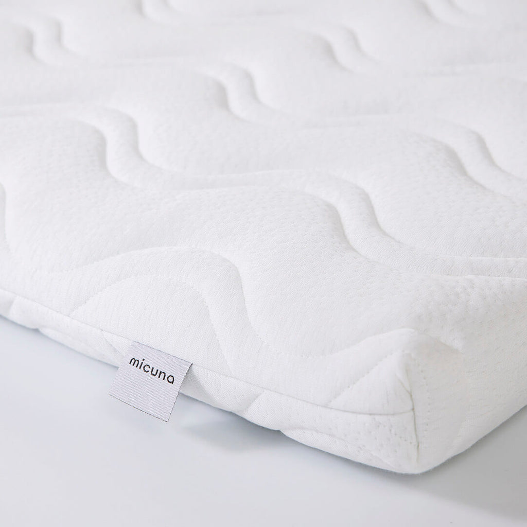 Detalhe do tecido branco acolchoado do colchão viscoelástico Micuna para mini-berço 80x50, com a etiqueta da marca visível.
