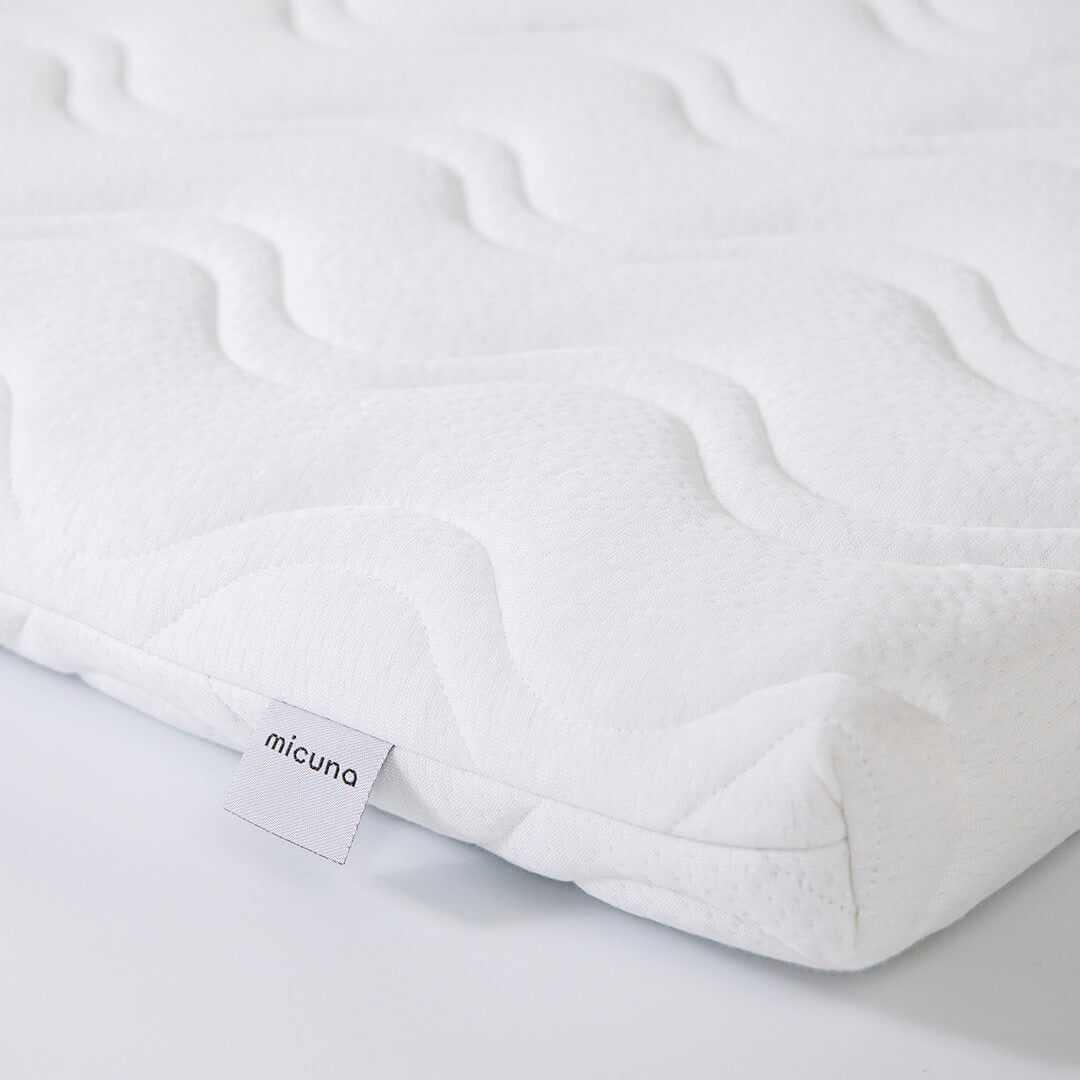 Detalhe do tecido branco acolchoado do colchão viscoelástico Micuna para mini-berço 80x50, com a etiqueta da marca visível.