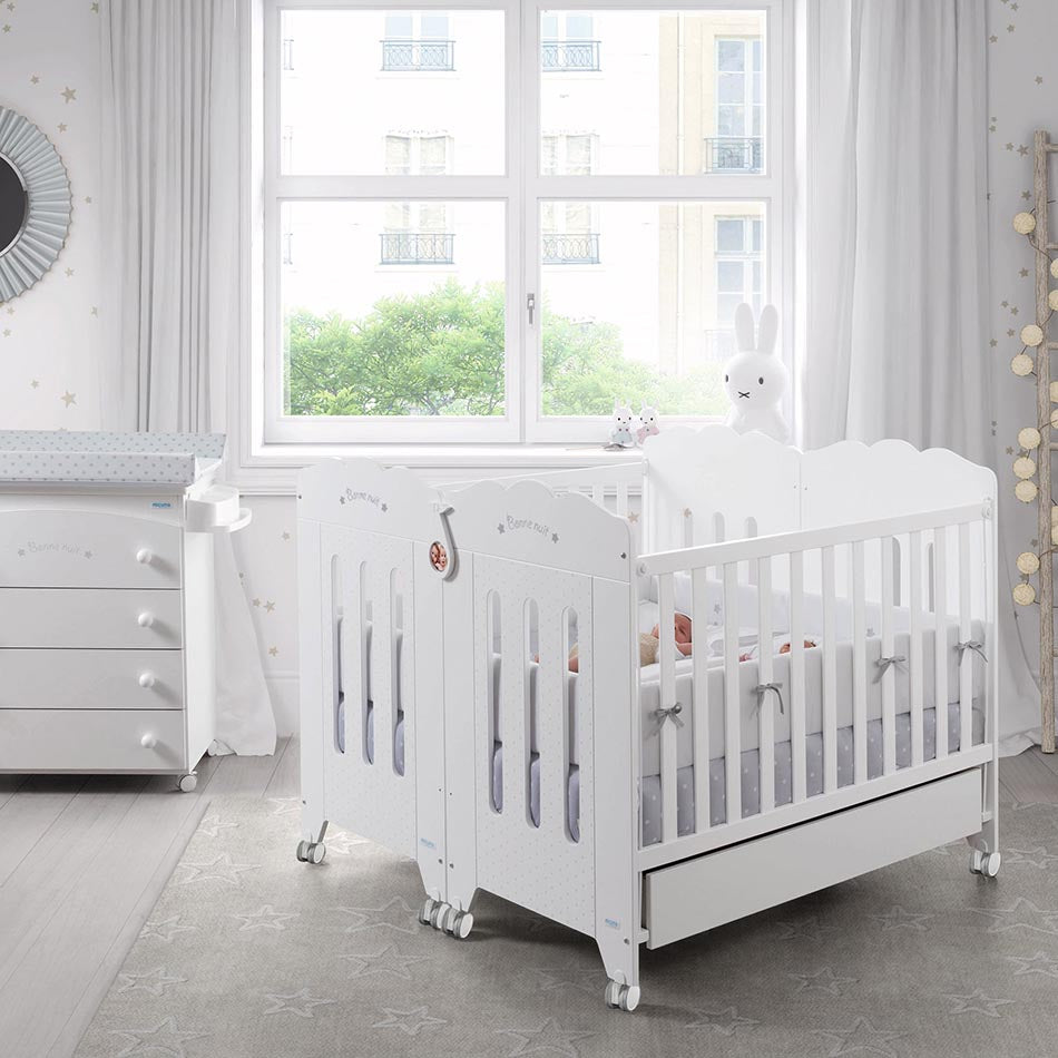 Quarto de bebé com duas camas Micuna brancas de 120x60 unidas pelo Kit Duo Gêmeos, com um bebé a dormir.