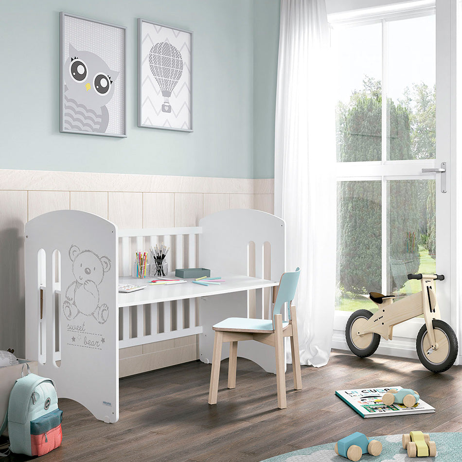 Um quarto infantil com um berço Micuna branco transformado em secretária, com uma pequena cadeira, materiais de desenho e brinquedos, junto a uma grande janela.