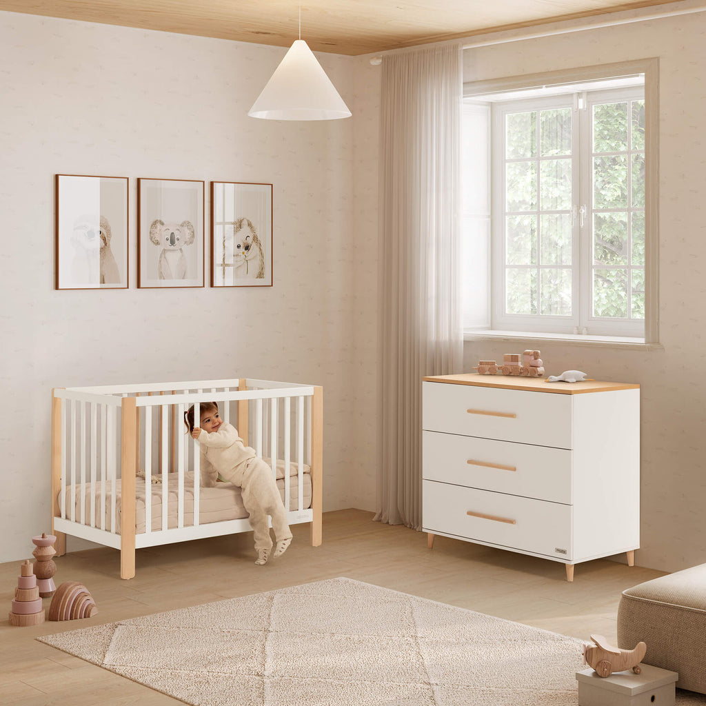 Berço Micuna Nayra de 120 x 60 em branco e madeira, com criança a sair, ao lado de uma cómoda a condizer num quarto infantil.