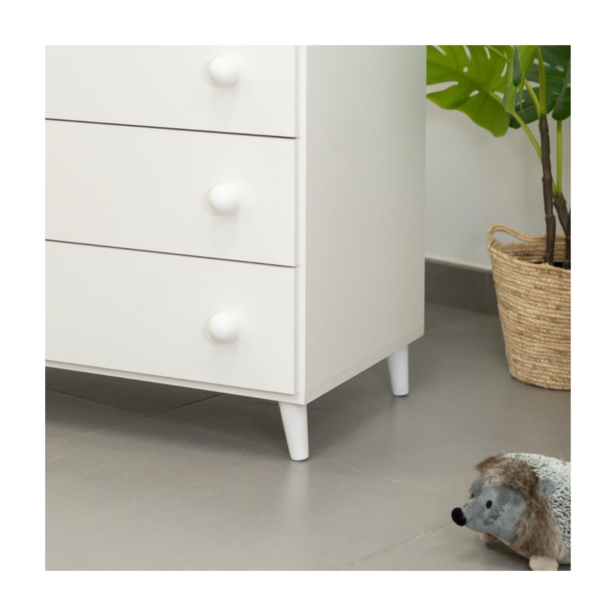 Detalhe da parte inferior de uma cómoda branca com pés de conversão brancos, ao lado de um peluche ouriço e uma planta em cesto.