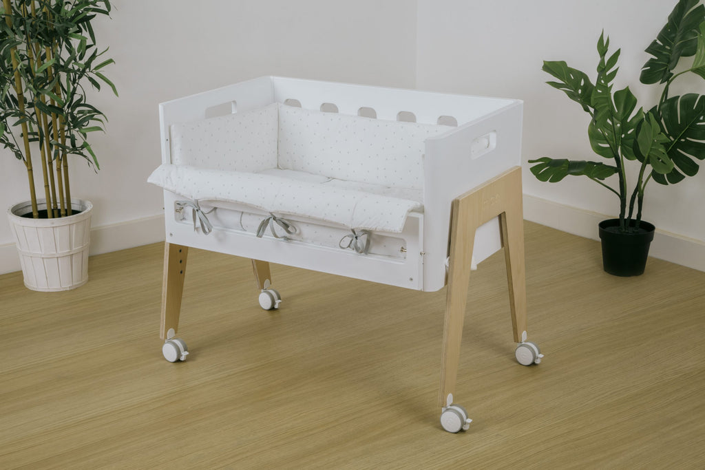 Minicuna branca com kit nórdico e protetor cinza claro de estrelas, em ambiente moderno com plantas.