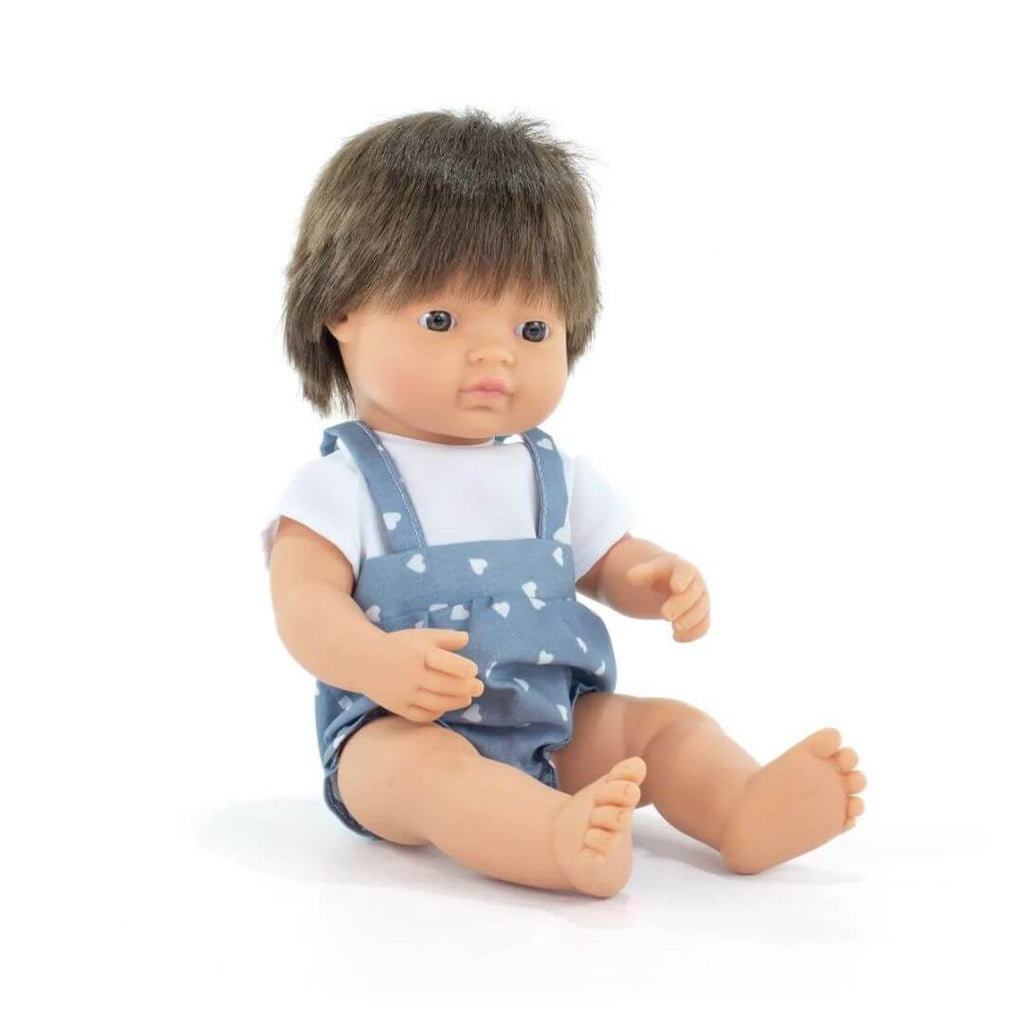 Boneco Miniland caucasiano de 38cm sentado, com cabelo castanho, vestindo jardineiras azuis com corações e t-shirt branca.