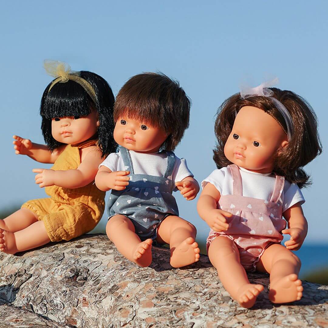 Três bonecos Miniland de 38cm (caucasiano, asiático e outro) sentados lado a lado ao ar livre, promovendo a diversidade e a inclusão.