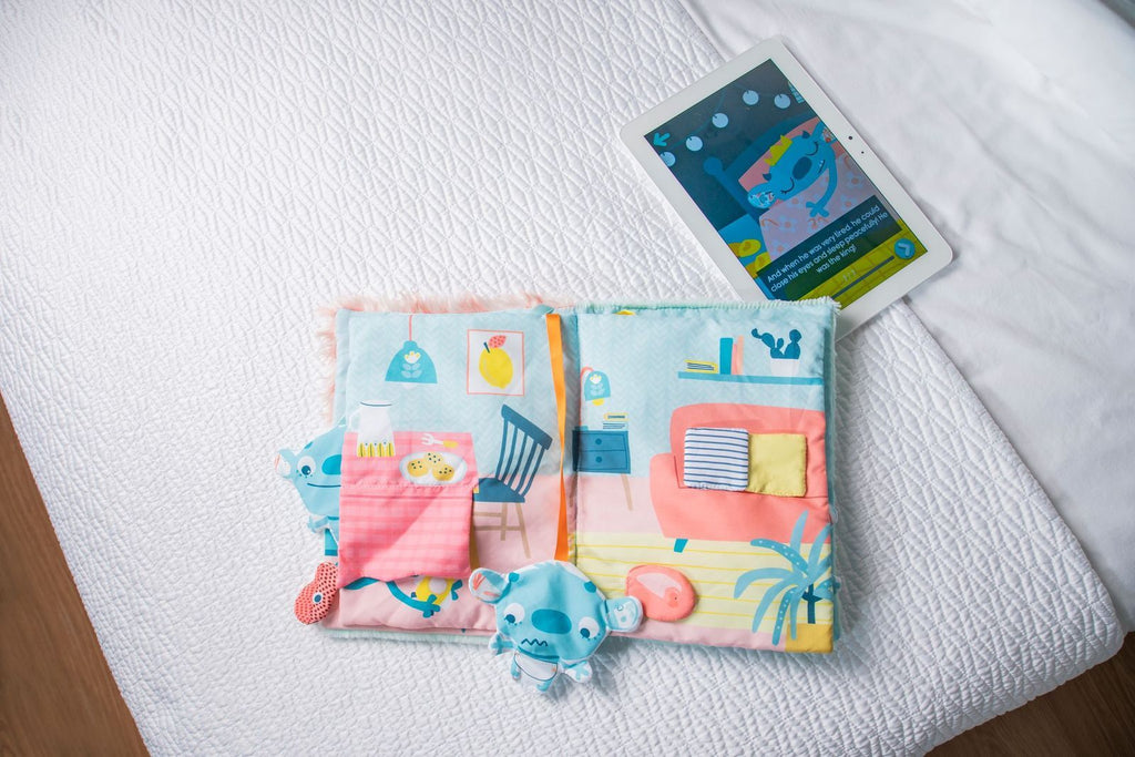 Miniland Livro sensorial Monstro aberto, mostrando páginas coloridas com ilustrações de casa e elementos interativos de feltro, ao lado de um tablet com a versão digital da história.