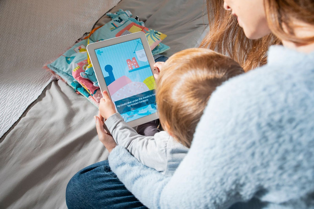 Mãos de um adulto e uma criança a segurar um tablet que exibe uma história digital, com o Miniland Livro sensorial Monstro desfocado em segundo plano.
