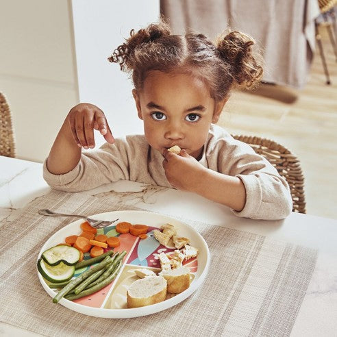 Criança pequena sentada à mesa a comer uma refeição diversificada no Prato NutriHealthy Miniland, que ajuda a separar os alimentos.