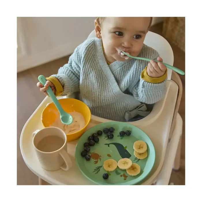 Bebé sorridente a comer papa com a colher do Miniland Conjunto de Alimentação 5 peças Ocean, com prato de baleia e taça amarela na cadeira de papa.
