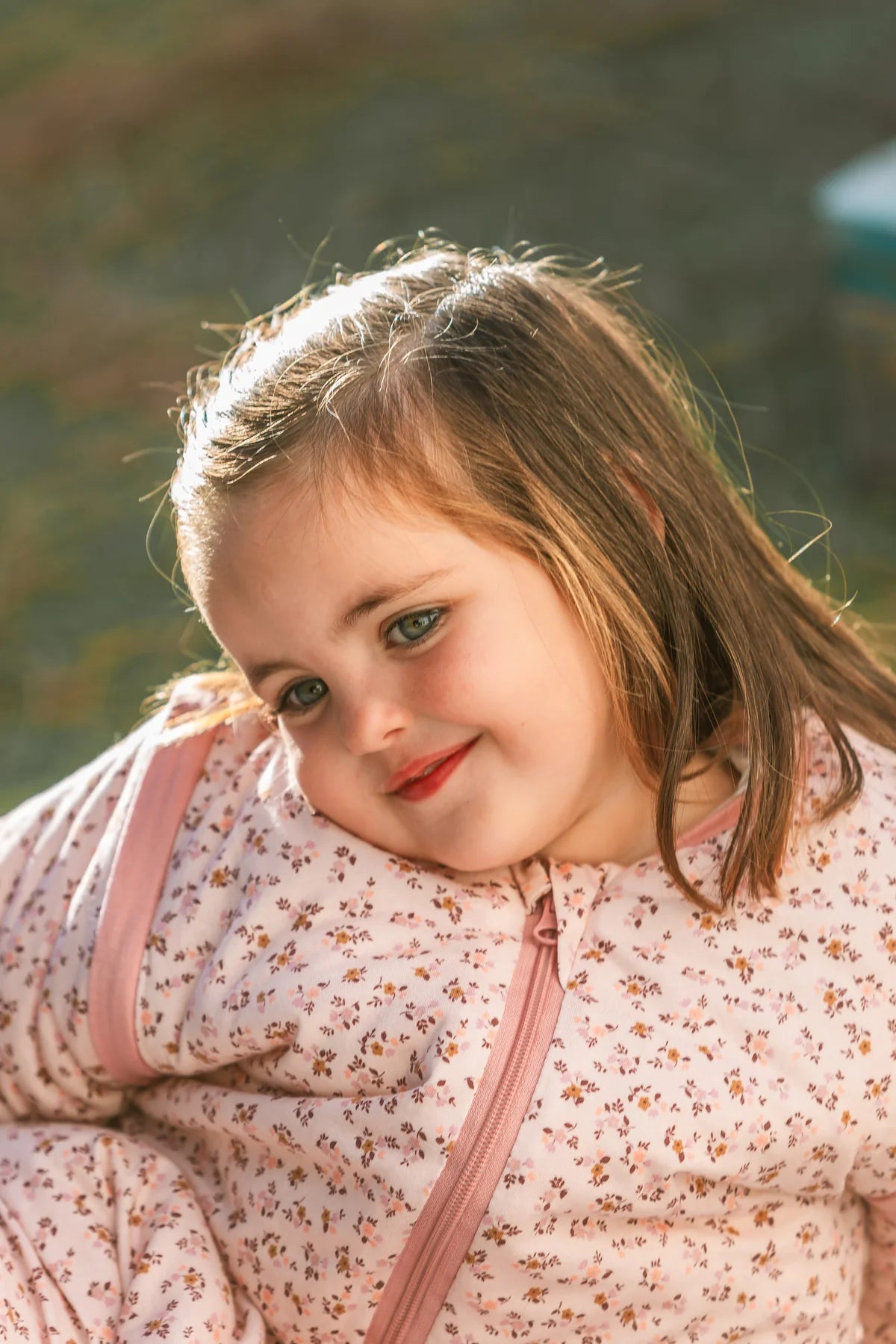 Retrato de uma menina sorridente com cabelos longos, a usar um saco de dormir MolisandCo TOG 2.5 com mangas e padrão floral rosa, à luz do sol.
