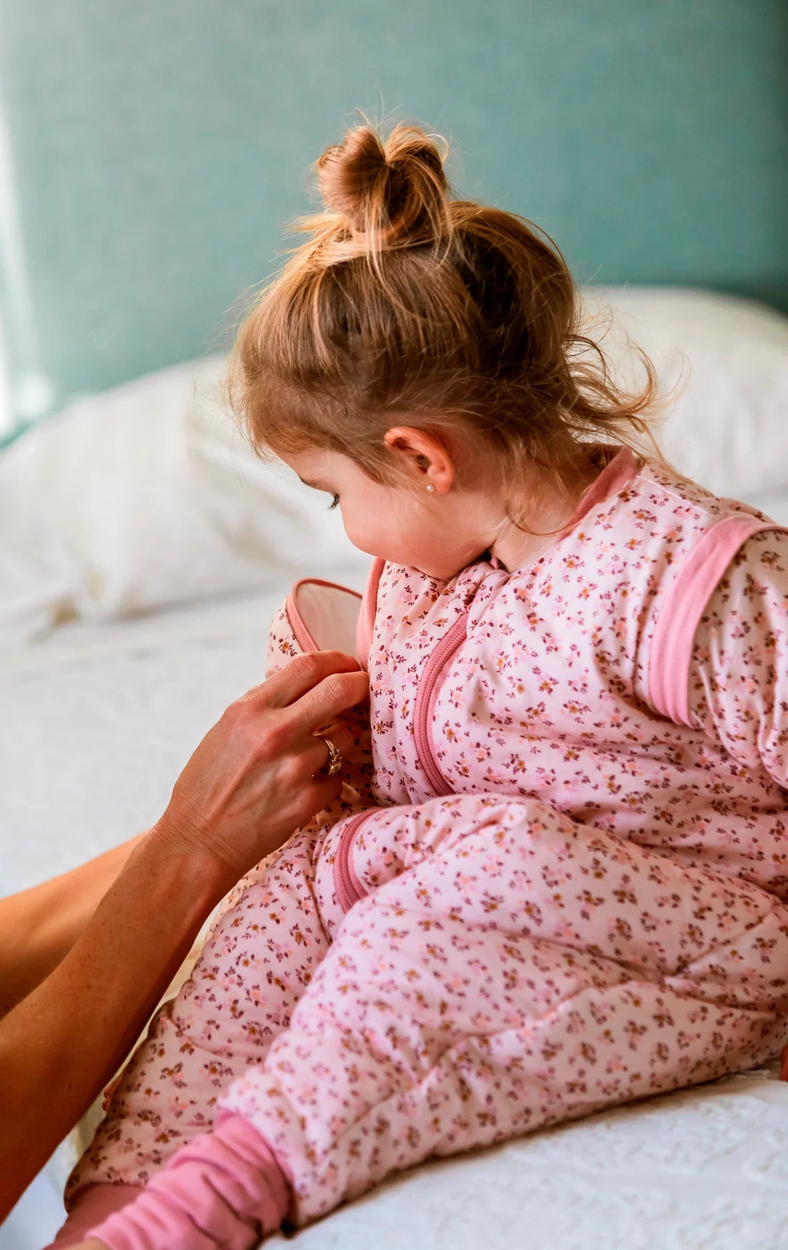Mãos de adulto a ajustar o saco de dormir MolisandCo TOG 2.5 Misty Rose num bebé sentado na cama, mostrando o fecho e o padrão floral.
