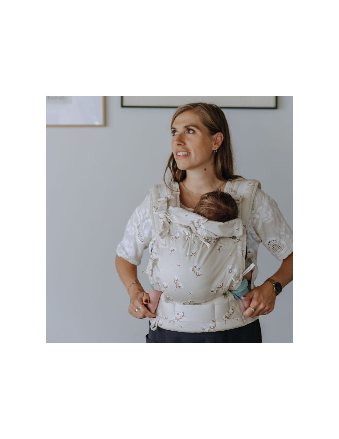 Mulher a usar uma blusa branca, carregando um bebé no porta-bebés Fidella Fusion Evolutiva Baby Cotton Flower Pale Grey. A imagem mostra uma visão de corpo inteiro, com o bebé posicionado de forma ergonómica e a mãe a olhar para a frente com uma expressão calma.