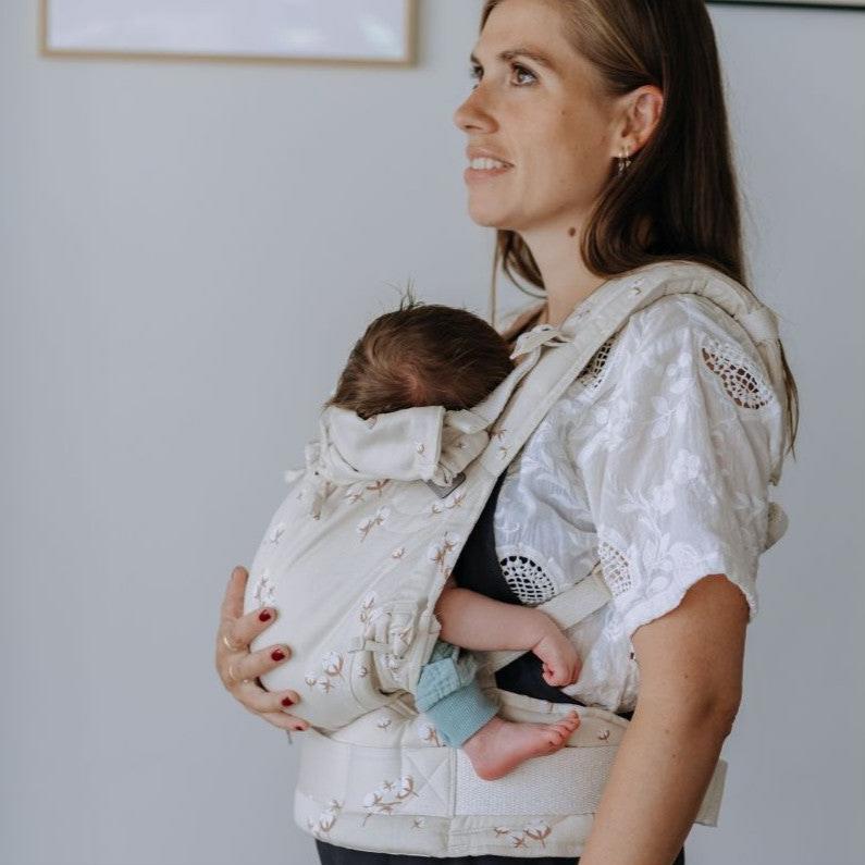 Mulher de pé com uma blusa branca, carregando um bebé num porta-bebés Fidella Fusion Evolutiva Baby Cotton Flower Pale Grey. O bebé está seguro e confortável no porta-bebés ajustado, com a cabeça encostada ao peito da mãe.