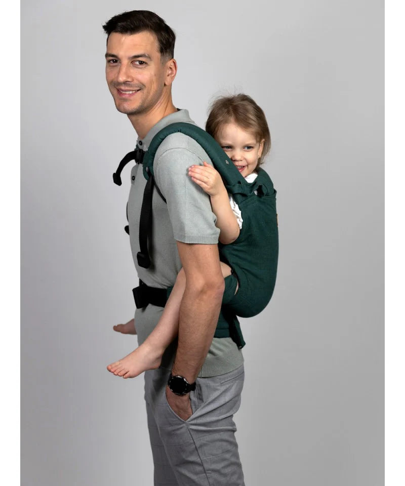 Vista lateral de um pai a carregar uma criança na Mochila Isara Elite Ajustável Evergreen Linen, numa posição de costas.