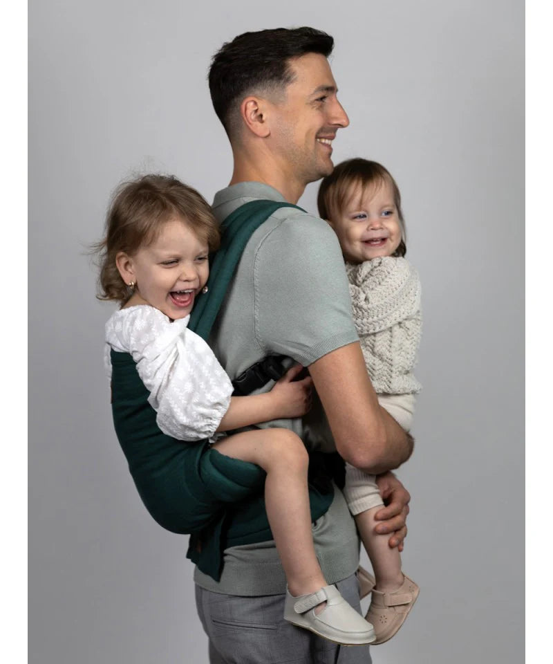 Um pai a carregar duas crianças pequenas na Mochila Isara Elite Ajustável Evergreen Linen, mostrando a capacidade de carga e o ajuste.