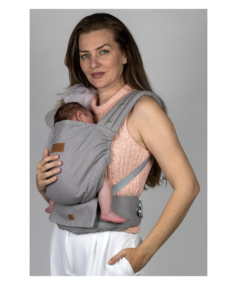 Mãe com cabelo comprido a usar a mochila Isara ELITE Ajustável na cor Misty Linen, transportando um bebé recém-nascido em posição frontal.